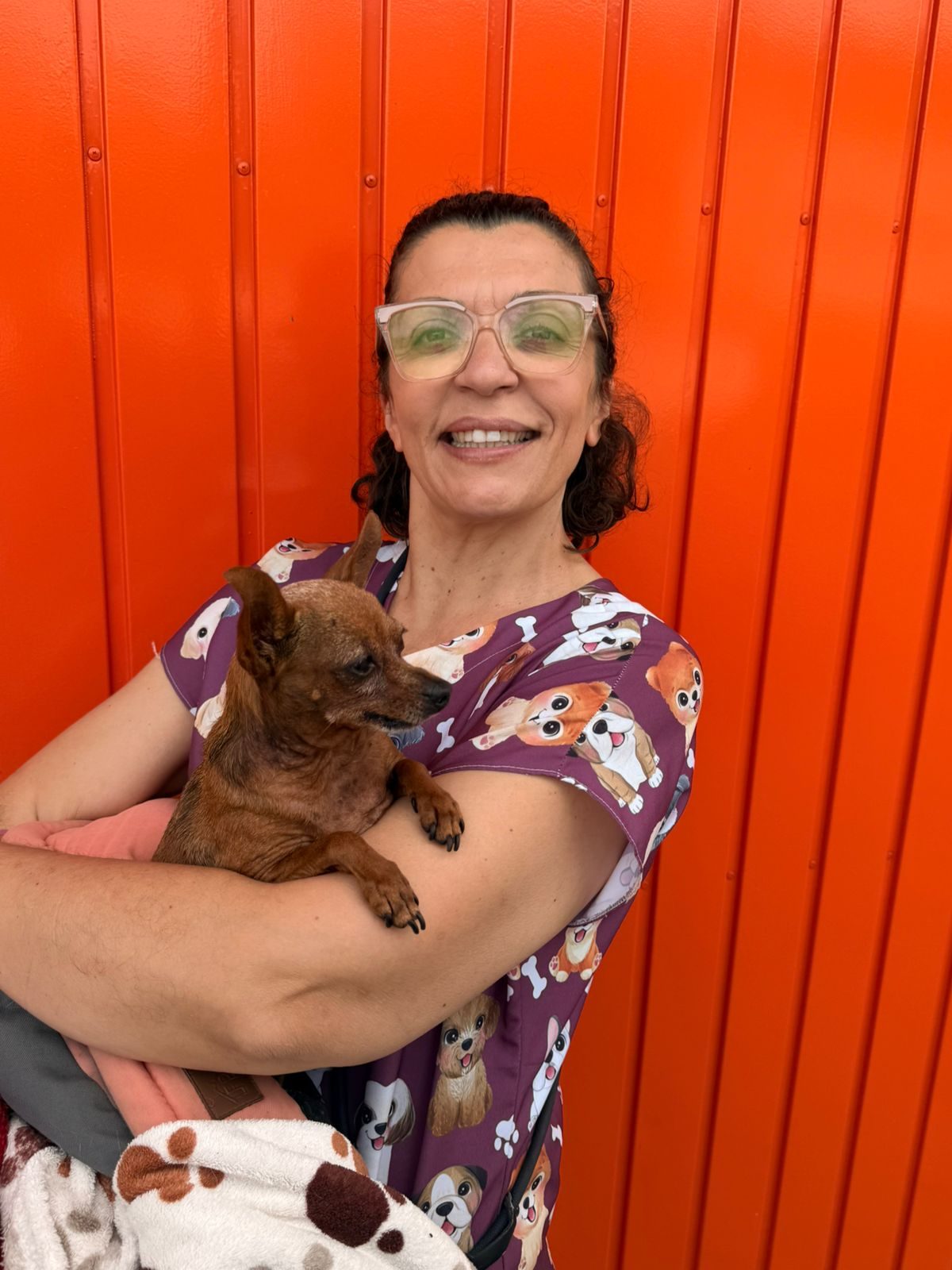 Dona Jô sorrindo com dois cachorros no colo, em frente ao portão laranja do hostel